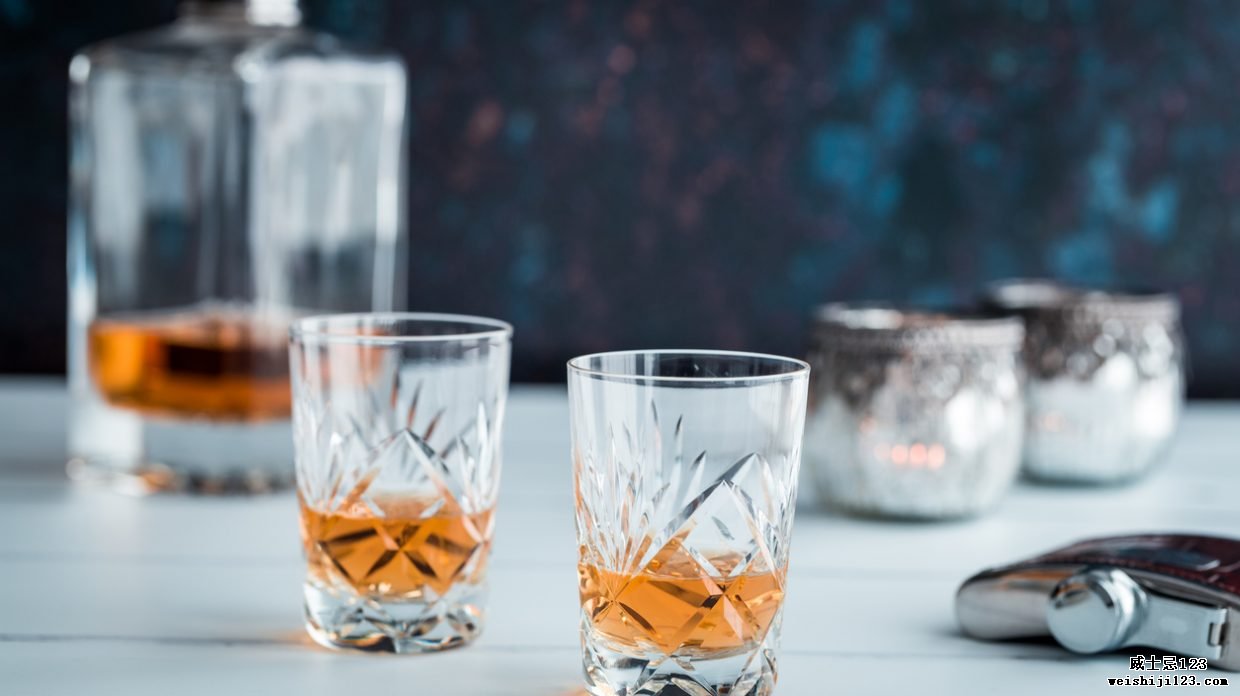 特写彩色图像，描绘了白色木质表面上的两杯精美水晶麦芽威士忌。 威士忌酒杯周围环绕着威士忌用具，例如玻璃醒酒器和酒壶