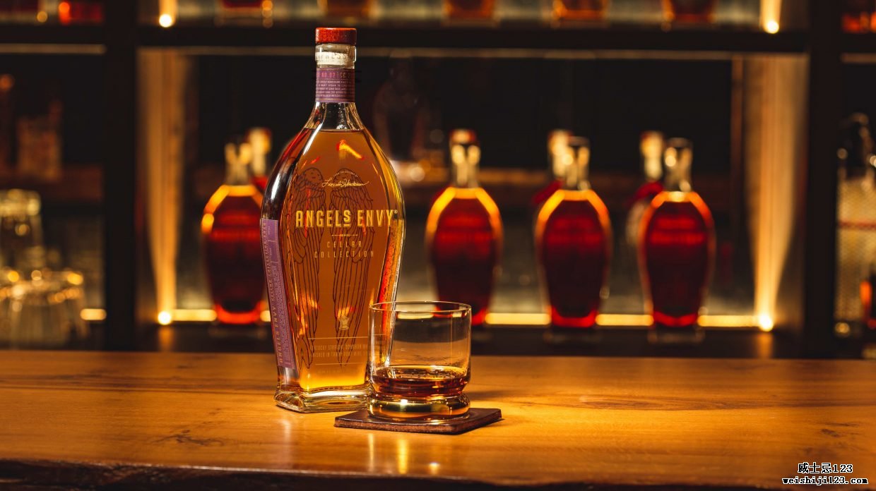 一瓶 Angel's Envy Tawny Port Cask-Finished Bourbon 在酒吧台上放着一杯威士忌和更多 Angel's Envy 酒瓶在背景中