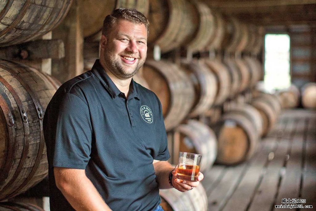 Jim Beam 酿酒大师 Freddie Noe 坐在一个侧放的威士忌酒桶上，左手拿着一杯威士忌，微笑着。