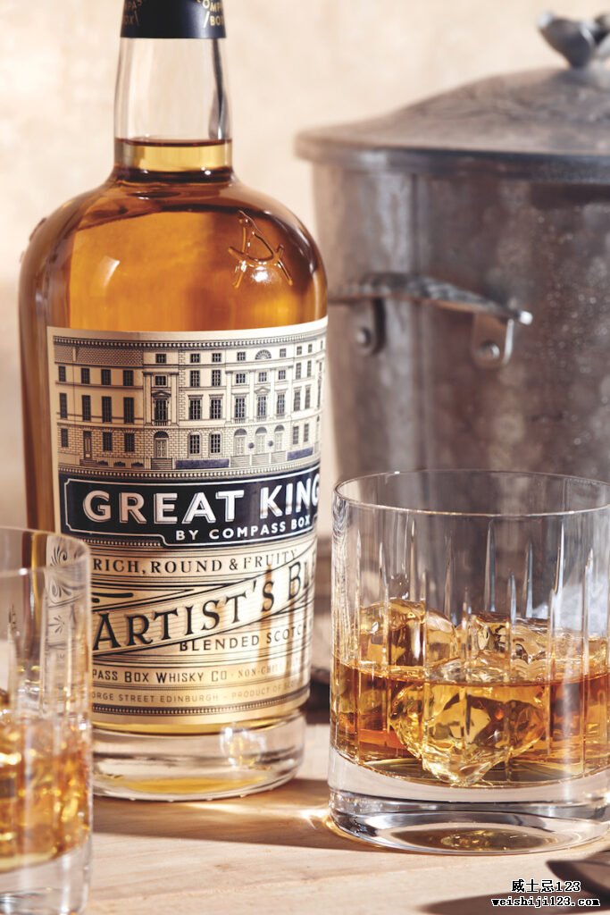 一瓶 Compass Box Great King Street Artist's Blend 放在木质表面上，还有一个酒吧勺和两个装有冰块和威士忌的岩石杯，背景是一个带盖子和小把手的金属冰桶冰桶.